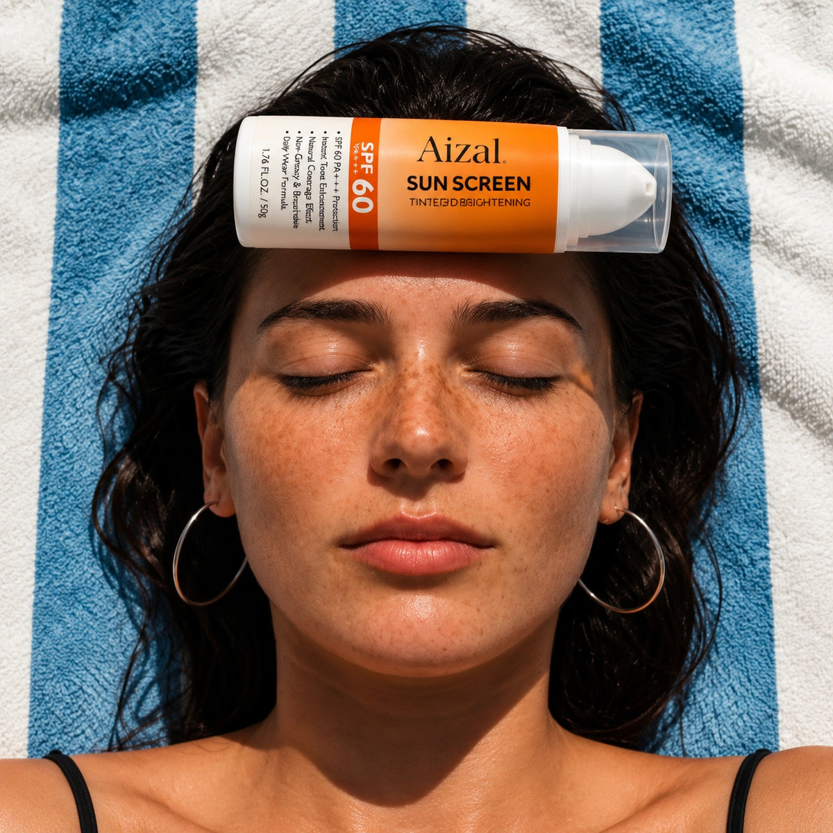 A woman lying on a blue and white striped towel with her eyes closed, wearing hoop earrings and having a tube of Aizal Sun Screen SPF 60 placed on her forehead.